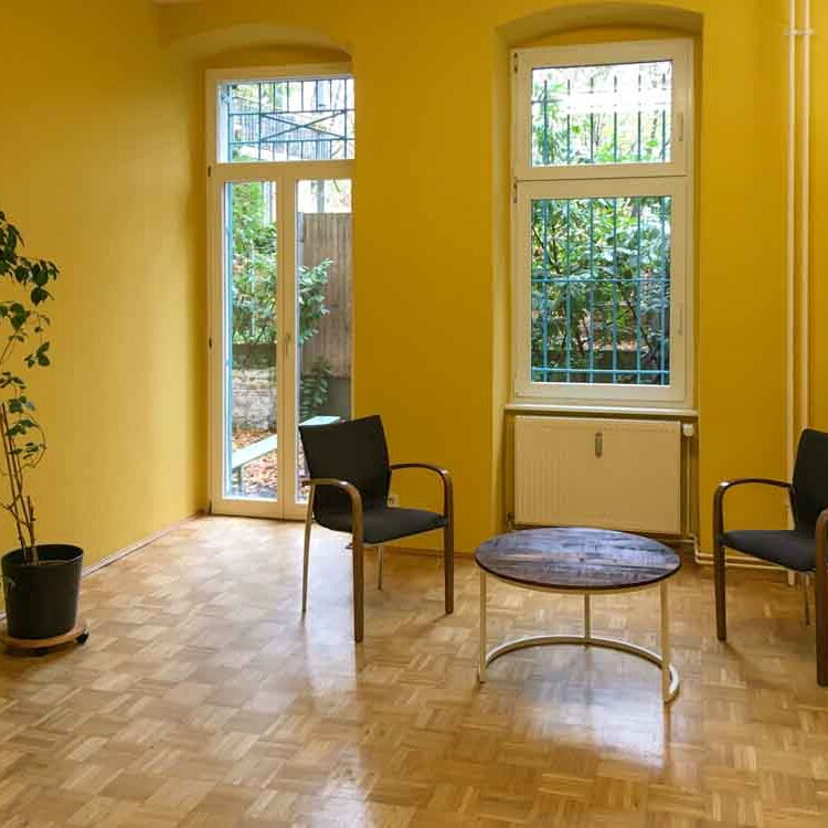 Der große Coachingraum mit einem runden Tisch und zwei Stühlen. An der Seite stehen zwei große Blumentöpfe. Die Wände sind gelb. Durch die Fenster ist ein Blick in den Garten möglich.